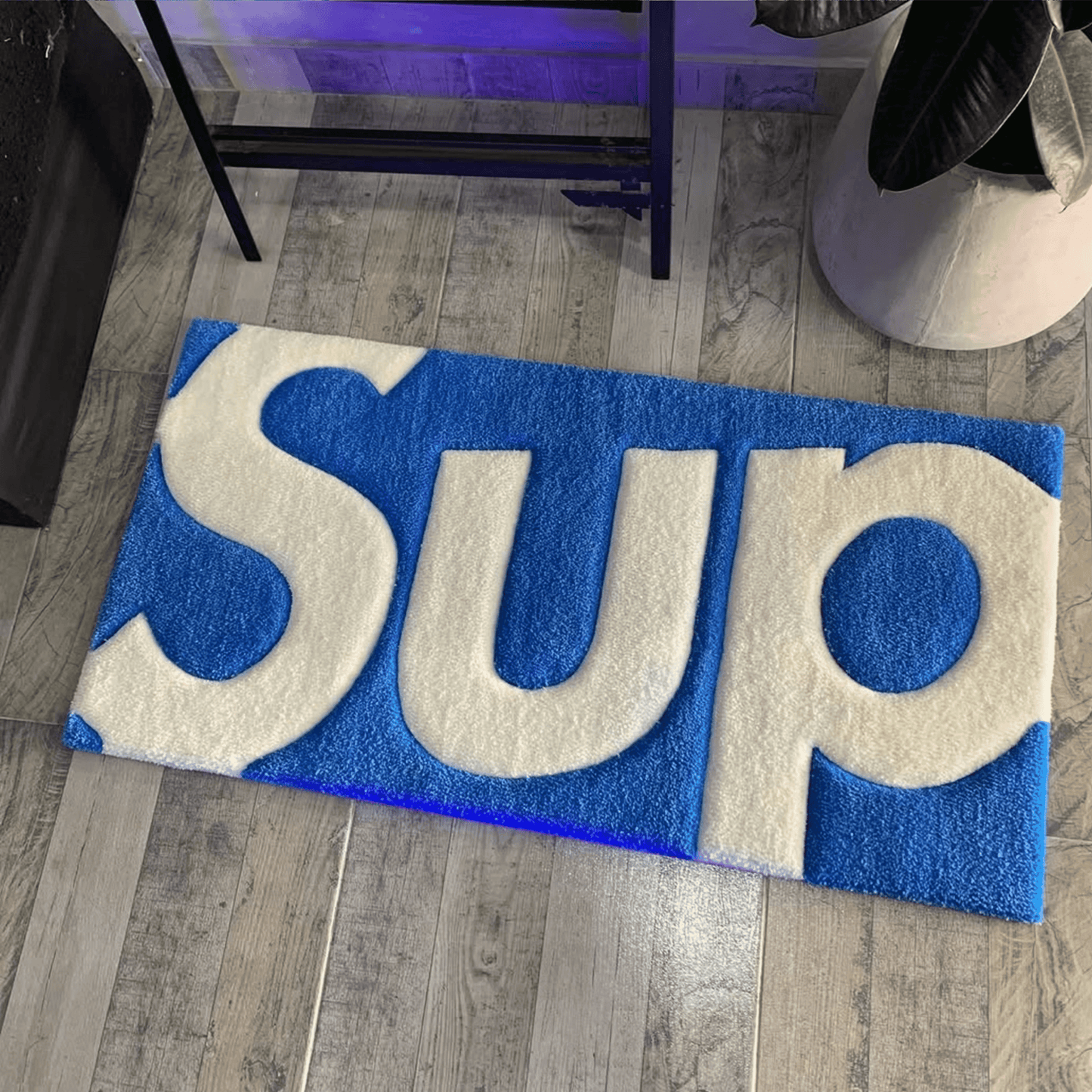 Supreme white blue box logo rug on wooden floor in bedroom. Handmade white blue tufted runner carpet. Sneakerhead hypebeast streetwear room floor decor.