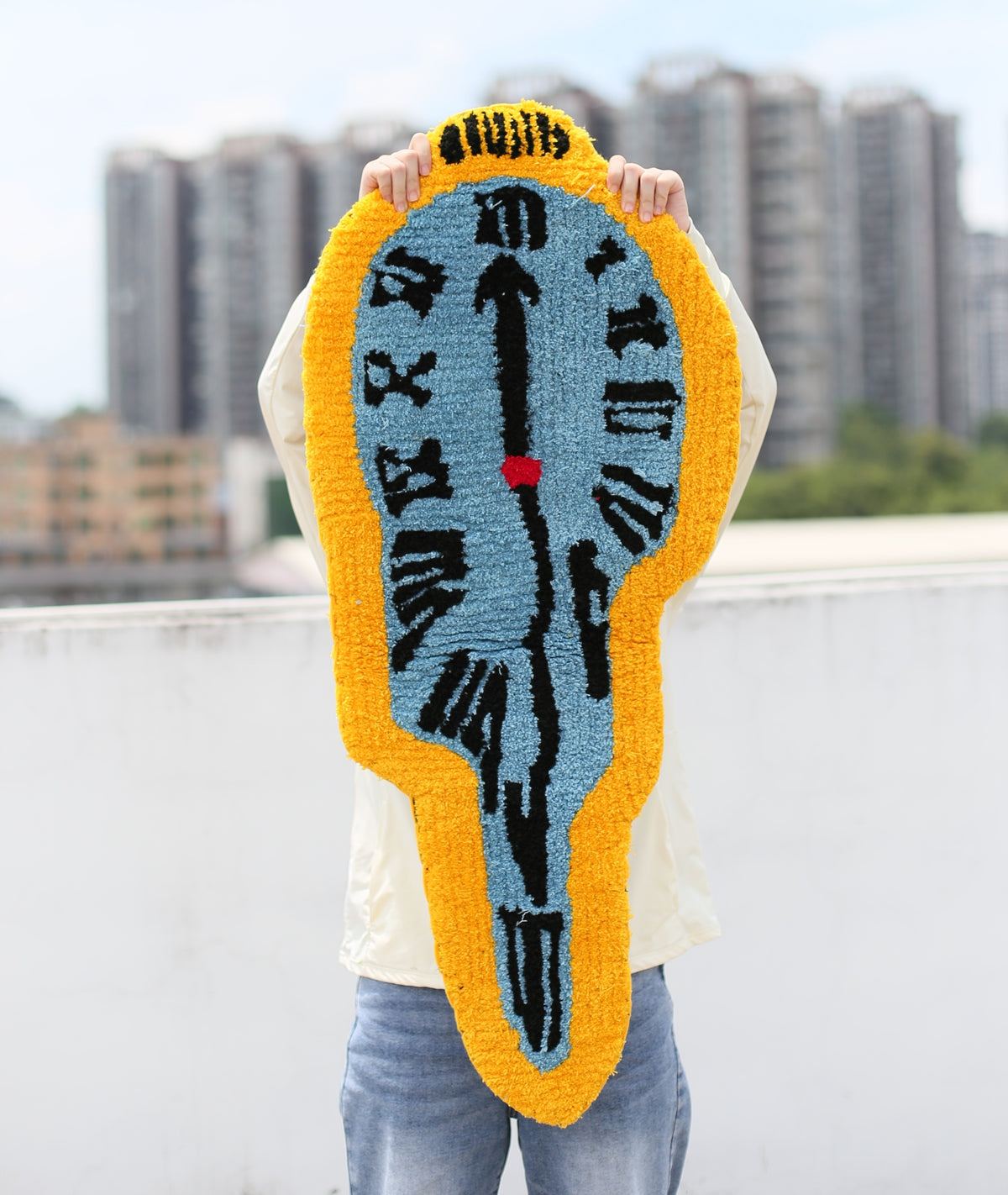 A person outdoors holds up a large, hand-tufted wall clock shaped rug or mat. The rug features a light blue face with black clock hands and Roman numerals, surrounded by a thick golden-yellow border, with a tiny red second hand.