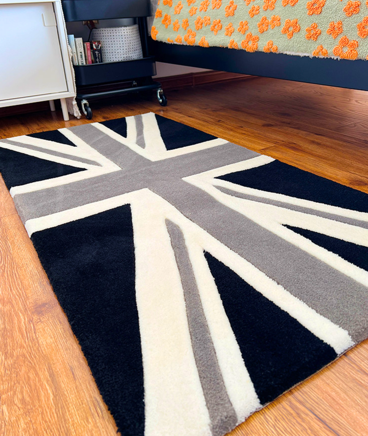 Rectangular flag rug beside a bed, thick tufted acrylic pile in off white and grey, angled view