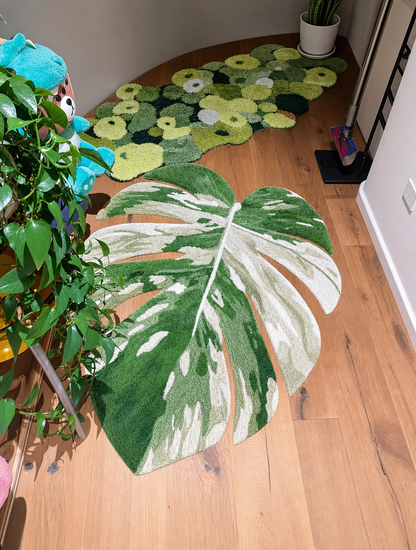 A large monstera leaf-shaped rug in green and white is placed on a light wood floor in a room, with other green patterned rugs visible in the background.