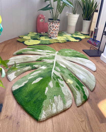 A large monstera leaf-shaped rug in shades of green and cream is placed on a wooden floor in a room setting, with other plants and decor visible in the background.