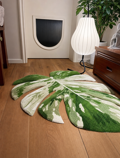 A large monstera leaf-shaped area rug in green and white sits on a light brown wooden floor in a modern room setting with a cat door, white floor lamp, and wooden furniture.
