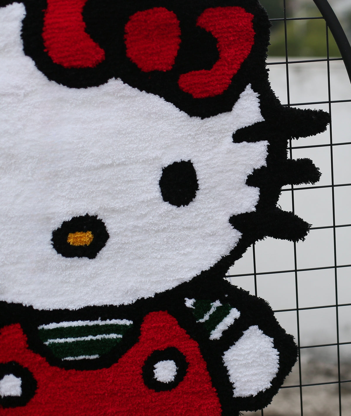 A close-up of a plush, looped textile product featuring Hello Kitty's face and upper body in red, white, black, and small yellow and green details, positioned against a black wire grid with an outdoor blurred background.