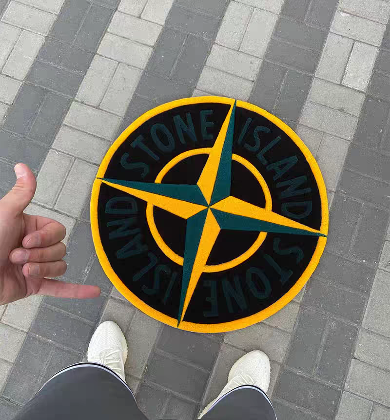 A Stone Island compass logo tufted rug on gray brick pavement, with a person's hand and shoes visible in the frame.