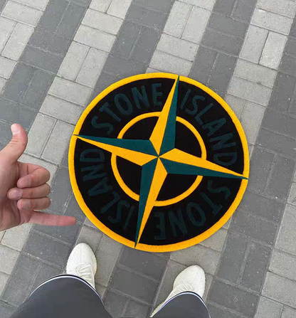 A Stone Island compass logo tufted rug on gray brick pavement, with a person's hand and shoes visible in the frame.