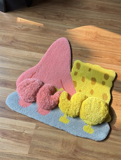 A plush, 3D-textured bath mat shaped like a pink sponge with yellow polka dots and pink bubbles, resting on a light blue base on a wooden floor.