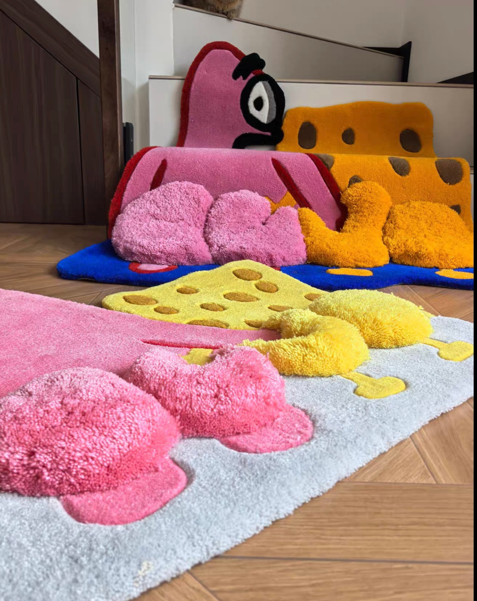 A set of plush, cheese-themed bath mats in pink, yellow, and orange, arranged on a light blue rug over a wooden floor.