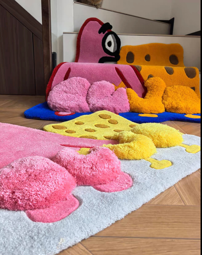 A set of plush, cheese-themed bath mats in pink, yellow, and orange, arranged on a light blue rug over a wooden floor.