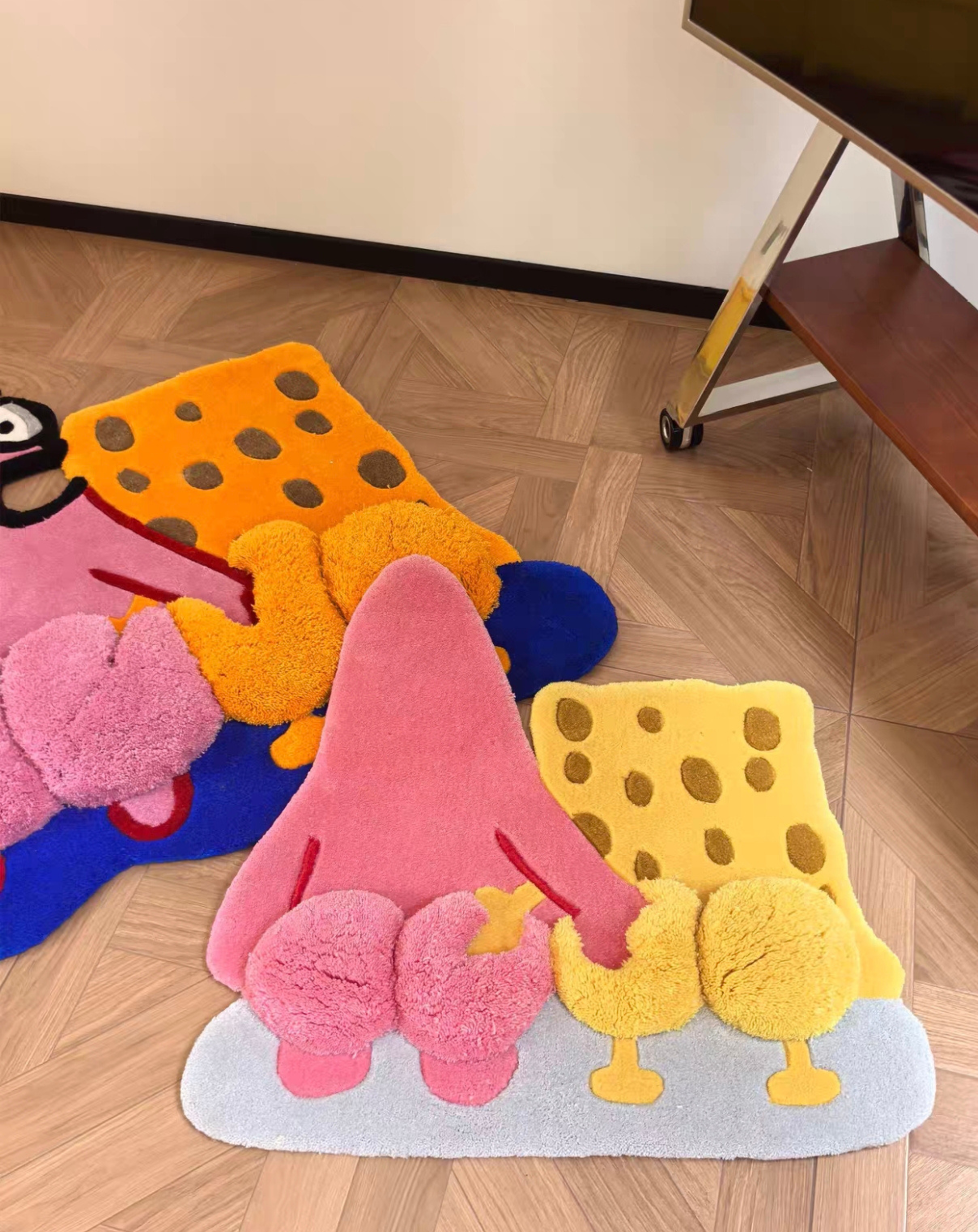 Three plush, cartoon-shaped bath mats in pink, yellow, orange, and blue are arranged on a wooden parquet floor in a room setting.