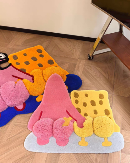 Three plush, cartoon-shaped bath mats in pink, yellow, orange, and blue are arranged on a wooden parquet floor in a room setting.