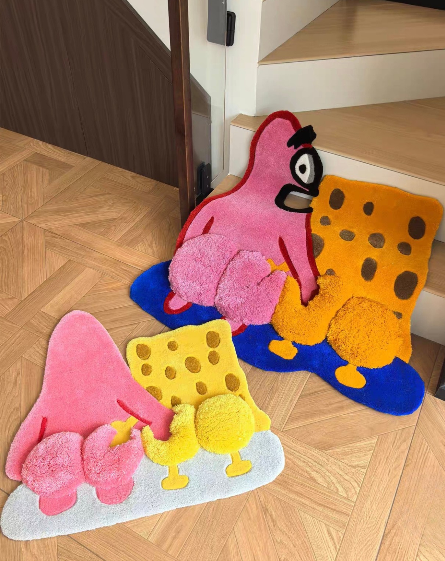 Two plush Patrick Star and SpongeBob-inspired floor mats in pink, yellow, orange, blue, and white, displayed on a light wood parquet floor near wooden stairs.