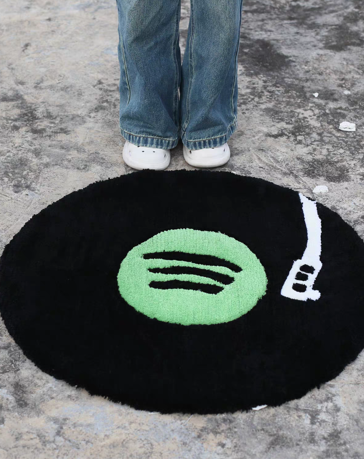 A round black plush rug shaped like a vinyl record, featuring a green Spotify logo in the center and a white stylized turntable detail, shown on a concrete floor with a person's legs and shoes visible above it.