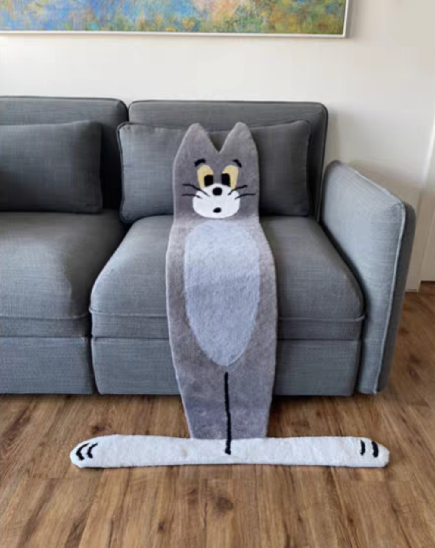 A plush gray cat-shaped floor mat with a cartoon face and long tail, placed in front of a gray sofa on a wood floor.