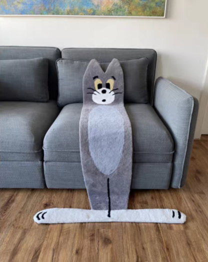 A plush gray cat-shaped floor mat with a cartoon face and long tail, placed in front of a gray sofa on a wood floor.