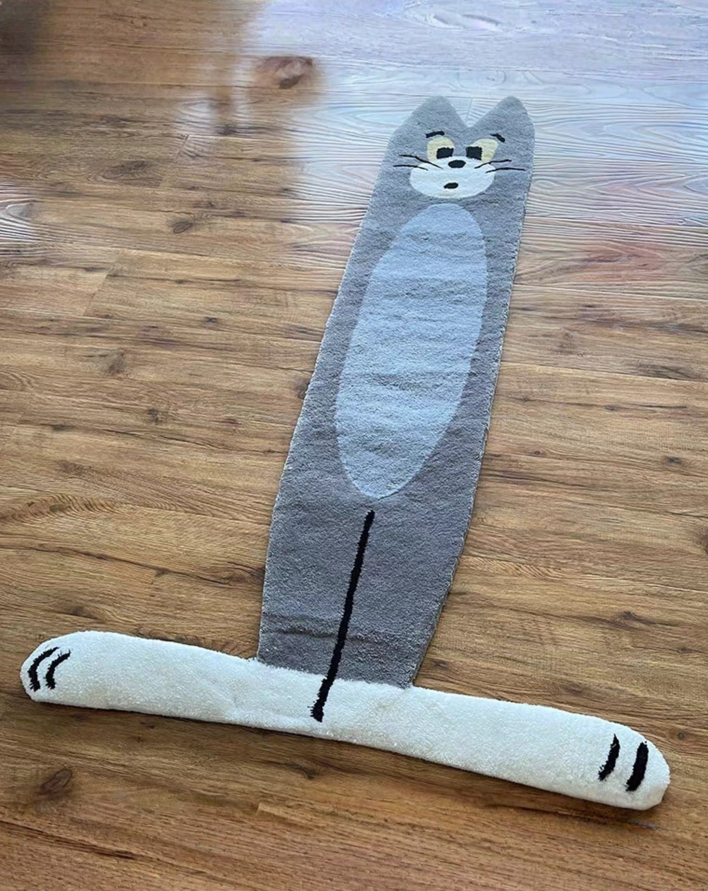 A novelty floor mat shaped like the character Tom Cat, featuring a gray body with a white oval belly, a cartoon face, and a long white tail with black paw prints, laid out on a wooden floor.